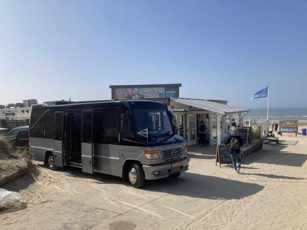 Uitvaartbus op het zand bij een strandpaviljoen met vlag